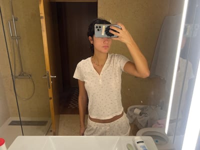 The scene is an interior view of a bathroom, located in Aldoar, Foz do Douro e Nevogilde, Portugal. A woman, captured from the chest to mid-thigh, stands centrally and takes a mirror selfie. She wears light-colored, short-sleeved pajamas featuring a small floral pattern, consisting of a V-neck henley top with buttons and matching shorts. A thin, dark beaded necklace is visible around her neck. Her right hand is raised, holding a smartphone (identified as an iPhone 14/15 Pro by its camera array) directly in front of her face, obscuring her facial features.
In the immediate foreground, a white sink basin is partially visible. On the counter to its left, a partial red cap is present. To its right, a white bottle labeled "Neutrogena 2 EM 1 Limpeza & Mascara" and a green applicator are positioned. The mirror itself incorporates two prominent vertical light strips along its right edge.
To the left of the woman's reflection, a modern shower area is visible, featuring a clear glass partition, beige tiled walls, a silver shower head, and a linear floor drain. Adjacent to the shower, a light-colored wooden door stands ajar, revealing a darker internal corridor with a patterned floor runner. To the right of the woman's reflection, a white toilet is visible, with a bidet faucet mounted on the adjacent wall. A pile of laundry, including a grey garment with a partially visible "ADIDAS" logo, rests on or near the toilet, alongside a roll of toilet paper. The bathroom walls are uniformly covered in beige-toned tiles or textured material.
