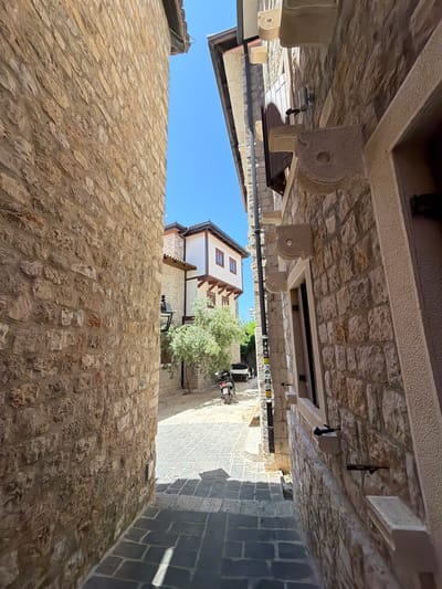 The image depicts a narrow cobblestone alleyway in the city of Ulcinj, Montenegro, flanked by two stone buildings. The left side of the alley is dominated by a rough-hewn stone wall, weathered and textured. The right side features a building constructed with similar stone, adorned with decorative stone brackets supporting a window ledge and a smaller window above. Shutters are visible on the upper window. The alleyway slopes gently downwards towards a bright, open area where a white building with dark-trimmed windows and wooden eaves is visible. A small, leafy tree stands near the white building, and a scooter is parked on the cobblestone street beyond it. The sky above is a clear, vibrant blue. The lighting suggests it is daytime, with shadows cast by the buildings and objects onto the alley floor. The scene conveys a sense of old-world charm and a typical Mediterranean streetscape.
