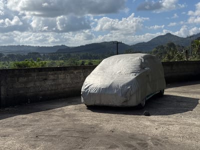 A silver car cover obscures a vehicle parked on a paved surface. Behind the car, a low, dark brick wall runs horizontally across the mid-ground, casting a shadow. Beyond the wall, a lush, green landscape with rice paddies and dense foliage stretches towards a series of rolling hills and mountains under a partly cloudy sky. The sky is a vibrant blue with dramatic white cumulus clouds. Electrical poles and wires are visible in the background, suggesting the presence of infrastructure. The overall impression is one of a car parked outdoors in a rural or semi-rural setting. The location is Sidemen, Indonesia.