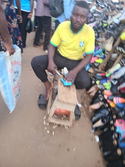 The image shows a person in a yellow t-shirt with a Brazilian flag emblem, sitting amongst a large collection of shoes and footwear. The setting appears to be an outdoor market, likely in Nyanya, Nigeria, given the context. The ground is sandy and appears to be a dry environment, suggesting a sunny day. The person is interacting with a small wooden structure and a blue object, possibly engaging in a repair or display activity. Various types of shoes are piled around, including sandals, sneakers, and boots, in a variety of colors. The overall impression is one of a busy marketplace, with the focus on the shoes and the person attending to them. There is a white bag with some text visible at the bottom of the image, but it is too blurry to be deciphered. The lighting suggests it is daytime.