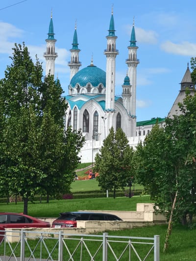 The image depicts a view of the Kul Sharif Mosque in Kazan, Russia. The mosque's prominent features include its white facade, turquoise domes, and tall, slender minarets topped with crescents. Lush green trees in the foreground partially obscure the mosque, with some branches and leaves creating a natural frame for the building.
In front of the mosque, a well-maintained lawn slopes gently upwards. A low, light-colored wall separates the foreground from the middle ground, where a red sedan and a red SUV are parked. The SUV has a license plate that reads "T243EP716". Beyond the cars and wall, the green lawn is visible, with some flowerbeds adding pops of color in the distance.
The sky above is a clear, bright blue with scattered white clouds, suggesting fair weather. The lighting indicates it is daytime, likely late morning or early afternoon, with the sun casting soft shadows. The scene is tranquil and serene, highlighting the architectural beauty of the mosque and its surrounding park-like environment. The overall impression is one of peaceful contemplation and architectural grandeur.