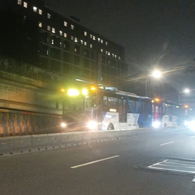 The image depicts a nighttime urban scene along a major transportation corridor in the Special Capital Region of Jakarta, Indonesia. In the foreground, an asphalt road surface is visible, partially marked with a faint white line. This road is separated by a concrete barrier from a dedicated busway. In the midground, multiple white and blue articulated TransJakarta buses are present on this busway, with their headlights and interior lights illuminated. The leading bus's headlights are prominently bright, and its destination sign area shows some orange illumination, though the text is unreadable. To the left of the busway, a multi-story, dark building with numerous uniformly spaced illuminated windows extends into the background. Adjacent to the busway and in front of the building, a long, vertical-striped wall, primarily yellow, blue, and brown in color, runs parallel to the bus lane. In the background to the right, bright streetlights are visible, casting light onto the scene. The overall environment indicates an active urban thoroughfare during non-daylight hours. The buses appear to be stationary or moving slowly within their designated lane.