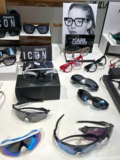 This image captures an indoor display of various eyeglasses and sunglasses, likely in an optical store or a fashion boutique in Koridallos, Greece. The scene is brightly lit, showcasing a range of eyewear styles on multiple shelves and hooks, suggesting products available for purchase.
The subjects include numerous pairs of glasses and sunglasses in different designs and colors. Some glasses are displayed on clear acrylic stands, while others are hanging from hooks, and a few are presented within their cases or boxes. Notable frames include classic dark-framed spectacles, stylish red and beige eyeglasses, and several pairs of sporty sunglasses with reflective or tinted lenses, some appearing to be shield-style.
Prominently featured is a black and white poster of a woman wearing glasses, next to which a black display bears the text "KARL LAGERFELD." Another brand, "DSQUARED2" with the word "ICON," is visible on black cases or pouches accompanying some of the sunglasses, indicating designer brands are part of the selection.
The overall activity is a retail display, designed to present eyewear attractively to potential customers. The environment appears clean and organized. The time of day is indeterminate, as it's an interior shot with artificial lighting. The image is notable for its clear focus on the diverse array of eyewear and visible branding.