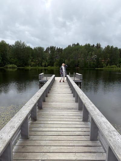 A person stands on a wooden pier next to a body of water. The person is wearing a patterned jacket, light-colored pants, and red shoes. They are facing towards the left of the frame with their right arm raised. The pier is made of light-colored wooden planks and has a railing. Stairs lead down from the pier into the water. The water is dark and appears still, with some lily pads visible. In the background, there is a dense forest of green trees. The sky is overcast with clouds, suggesting a cloudy or possibly rainy day. The overall scene is serene and natural. The location is indicated as Mars, Russia. No text is visible in the image.