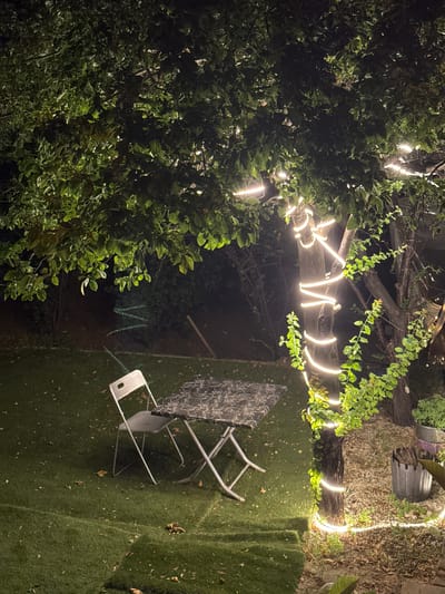 The image depicts a dimly lit outdoor scene, possibly a backyard or patio, at night. A large tree dominates the foreground, its trunk wrapped in a string of warm white LED lights that create a luminous spiral effect. The tree's branches, laden with green leaves, extend overhead, some of which are also illuminated by the lights. On the artificial grass-covered ground, a foldable metal table with a patterned top and a matching foldable metal chair are positioned. The table and chair appear to be set up for outdoor seating. Further in the background, a zigzag pattern of green lights is visible, possibly another decorative lighting element. The surrounding area is dark, with hints of foliage and possibly other trees or structures. The scene is quiet and serene, suggesting a peaceful evening atmosphere. There are no people present in the image. The location is identified as being in Şişli, Turkey.
