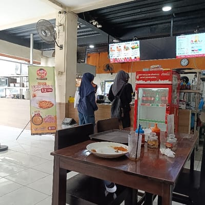 The scene is a food stall in Kaliwungu, Indonesia, located at coordinates -6.962, 110.256. There are two people in the scene. One person is a woman standing in front of a food stall. The woman is wearing a blue headscarf and a blue shirt with jeans. The other person is a woman standing in front of a red cooler. The woman is wearing a black headscarf and a long-sleeved black shirt with black pants. The stall features a large red cooler that is stocked with beverages and sauces. There is a large poster for "Telur Crispy" with prices listed and a smaller poster with details of the food. There is a wooden table with a plate with the remnants of food and a glass of water sitting on it. There is a stool beside the table. The room is decorated with brown and yellow tiles on the wall, and a brown ceiling.