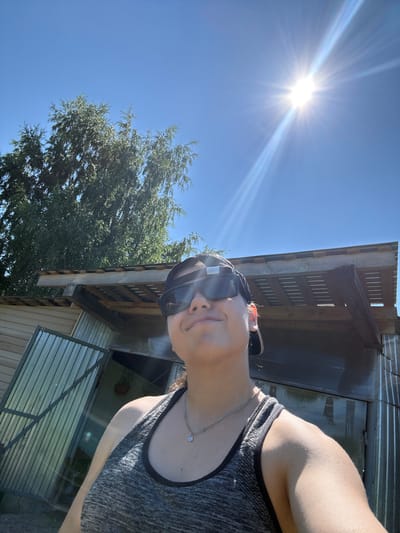 In a low-angle, wide shot, a person is visible from the chest up, taking a selfie outdoors on a bright, sunny day. The person is wearing a gray tank top, a silver necklace with a clover pendant, sunglasses, and a black baseball cap. They are smiling slightly and looking upwards towards the sun. In the background, a large tree with green foliage is on the left, and a rustic structure with a corrugated metal exterior and a wooden roof is on the right. The sky is a clear, bright blue, with the sun prominently featured in the upper right quadrant, creating lens flares and starburst effects. The overall environment suggests a rural or suburban setting, possibly a backyard or a recreational area. The image does not provide specific clues to pinpoint the exact location within the city of Elovo, Russia, beyond the general description of an outdoor scene with a building and vegetation.