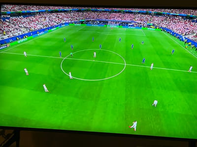 This is a high-angle, full-frame, medium shot of a professional soccer match on a brightly lit, green grass field. The field is marked with white lines, including a center circle. Spectators in red and white attire fill the stadium seating that surrounds the field. Several players, dressed in blue and white uniforms, are scattered across the field, some in motion. A large, white banner with green text that reads "aramco" runs along the perimeter of the field. The stadium appears to be modern and well-maintained. There is no indication of the city of Florești, Romania in this image.