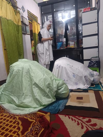 The image depicts an indoor scene, likely a home, in Cianjur, Indonesia. Two individuals are engaged in prayer, a common practice in Muslim households. One person, draped in a light green prayer garment, is in a kneeling or prostrating position, their head covered. Another individual, wearing a white hijab and a long white garment, stands upright, holding a mobile phone in their hands, possibly engaged in religious recitation or consultation. They are positioned in front of a tall, dark glass-fronted cabinet filled with various items, including books and decorative objects. To the right of the cabinet are white plastic drawers, commonly used for storage. The room is decorated with colorful rugs on the floor, one of which is predominantly red with intricate gold patterns. Green and yellow curtains hang on the left side of the room, partially obscuring a window. The overall impression is one of a domestic, religious observance.