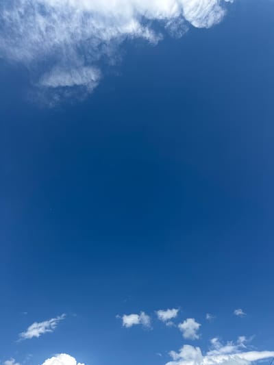 This is a full-frame, low-angle shot of a vast, clear blue sky. The dominant subject is the sky itself, with a few scattered white clouds. On the left side of the frame, a large, wispy cloud formation stretches from the bottom edge upwards, its texture appearing soft and feathery. Towards the right edge, several smaller, puffier clouds are visible, some appearing as distinct, isolated masses, while others are more elongated and streaky. A very faint, thin line, possibly a power line, is visible at the very top right edge of the frame. The lighting suggests it is daytime, with the bright blue sky and white clouds indicating sunny weather. There is no text visible in the image, and no people or objects other than the clouds are present. The overall impression is one of a bright, clear day. The location context of Каменномостский, Russia, is not visually apparent from the image itself.