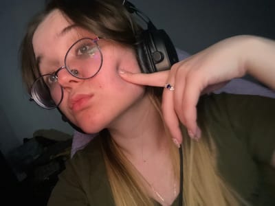 The image depicts a female individual in an indoor setting. The specific location in Pushchino, Russia, cannot be determined from the visual information provided. The subject has long, light-colored hair and wears round spectacles with dark frames and a septum piercing. Black over-ear headphones are worn. She is clad in an olive green upper garment, possibly over a light purple layer, and a thin silver-colored necklace. Her left hand is raised, with the index finger pressing into her left cheek, creating a distinct indentation. A ring with a dark blue oval stone flanked by smaller clear stones is visible on her left ring finger. Her fingernails are manicured with a glitter-detailed polish. The background is dark and out of focus, showing indistinct shapes in the lower left portion of the frame. The subject looks directly towards the viewer, illuminated by a frontal light source.