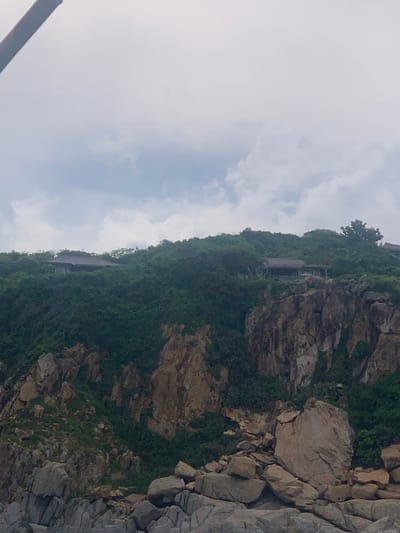 The image shows a cloudy sky over a rugged, vegetated cliff face with a few buildings nestled among the trees near the summit. The cliff is composed of large rock formations, some of which have tumbled down to form a rocky base. The buildings appear to be houses or villas, with dark, sloped roofs and some visible windows. The vegetation is dense and green, covering the hillsides. The scene is likely a coastal landscape.