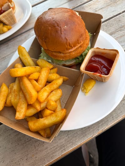 A close-up, top-down view of a burger and fries meal is presented on a white plate, situated on a wooden table. The meal includes a burger with a golden-brown bun, lettuce, and a crispy patty, served in a brown cardboard container. Alongside the burger are thick-cut fries, also in a brown cardboard container, with a small waffle cone cup filled with ketchup. A single fry is on the plate next to the ketchup cup. The wooden table has a weathered, grey finish. The background shows a blurred white plate with additional food items, including a waffle cone. The scene is well-lit, suggesting an outdoor or well-lit indoor setting. No individuals are visible in the image, and there are no discernible landmarks or contextual clues to pinpoint a specific location within Lörrach, Germany.