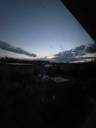 The image depicts a rural scene at dusk in the city of Burenka, Russia. The sky is a gradient of blue and gray with some scattered clouds, and a hint of pinkish-orange on the horizon suggests the setting sun. In the foreground, a dark, cluttered yard is visible. Various objects are strewn about, including what appears to be a tire and some coiled cables. Several outbuildings or sheds are present, with one in the mid-ground having a corrugated metal roof. A dark-colored vehicle is parked near one of the structures. The overall impression is one of a utilitarian, somewhat unkempt outdoor space in a rural or semi-rural setting.