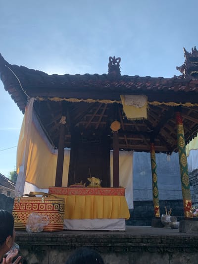 The image captures a low-angle view of an outdoor altar or shrine, likely in Sukasada, Indonesia, bathed in the warm glow of sunlight against a clear blue sky. The altar is covered by a dark, tiered roof with ornate carvings and is supported by decorative pillars. Yellow and white fabrics drape the sides of the structure, adding a touch of color. In the foreground, a person's head and shoulder are visible, holding a clear plastic cup. To the left, a woven basket with red and yellow patterns sits on a stone ledge, next to a plastic-wrapped bundle. The altar itself features a red and gold patterned border with a yellow fabric skirt. Several offerings, including what appear to be small incense sticks and decorative items, are arranged on top of the altar. In the background, a textured gray wall and the roof of a neighboring building are visible.
