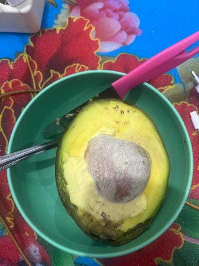 This image captures a close-up view of a halved avocado with its large pit resting in a vibrant green bowl. A pink-handled knife is positioned on the left side of the bowl, with its blade subtly touching the avocado's flesh. A spoon is visible at the bottom of the bowl, partially submerged and angled.

The avocado is ripe, with bright yellow flesh and a slightly darker green peel. Small brown specks are scattered across the flesh, suggesting ripeness. The pit is light brown and rough in texture.

The bowl sits on a colorful tablecloth or placemat featuring a bold pattern of red and pink floral designs outlined in yellow against a blue background. In the top left corner of the image, a white charging cable and adapter are visible, hinting at an electronic device nearby.

The lighting appears to be artificial and overhead, creating some subtle shadows within the bowl. The overall scene suggests a casual setting, likely indoors, possibly in a kitchen or dining area. No people are visible in the frame. The image is well-lit, and the focus is on the avocado and the surrounding items. The colors are rich and saturated, especially the greens of the bowl and avocado, and the reds and blues of the patterned surface. The image does not contain any visible text.