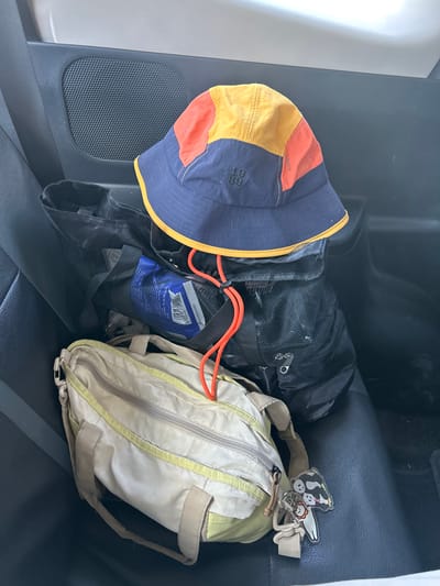 A bucket hat with blue, orange, and yellow panels sits atop a stack of bags inside a vehicle. The hat has the numbers "19 89" embroidered on it. Below the hat are two black mesh bags, one containing a blue bag with white lettering. In front of these is a light-colored shoulder bag with green trim and a zipper closure. A keychain with cartoon animal figures is attached to the bag. The scene is shot from a slightly elevated perspective, looking down into the back seat of a car. The interior of the car is dark, with the black leather seats visible. The car appears to be parked, possibly in the city of Kalabbirang, Indonesia.