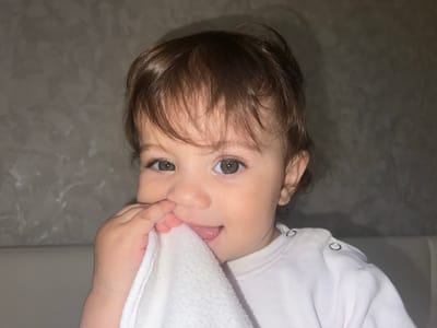 A close-up shot of a baby with brown hair and hazel eyes, holding a white cloth in their left hand and partially putting it in their mouth, sticking their tongue out. The baby is wearing a white long-sleeved shirt with snaps on the shoulder. The background is a neutral gray, resembling a wall texture. The location is not discernible and is unlikely to be Elena, Bulgaria, based on the visual information provided.