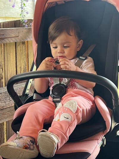 A baby girl is secured in a pink baby stroller, possibly outdoors. She is wearing a pink top, pink pants with fox details on the knees, and patterned shoes. The stroller has a black frame with a pink interior. The stroller appears to be near a wooden structure, with a glimpse of purple flowers in the background. The image may have been taken in Elena, Bulgaria, but there is no direct evidence.
