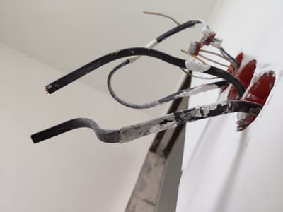 The image shows a close-up of electrical wires being installed in a wall. The scene is an indoor setting, likely a residential or commercial construction site. Several black electrical cables are visible, some with exposed copper wire ends. Some of the cables are wrapped with white tape, and there are red plastic junction boxes protruding from a hole in the white wall. Red electrical connectors are also visible, with wires of various colors (blue, yellow, and white) entering them. The overall impression is one of unfinished electrical work. There is no visible text in the image, and no cues about the time of day or weather.