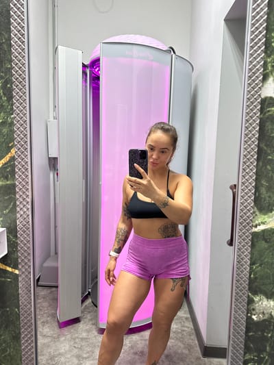 A young woman with dark hair tied back in a bun is taking a selfie in a tanning salon. She is wearing a black sports bra and short purple shorts. She has tattoos on her arms and torso. The tanning bed is visible behind her, with pink lights glowing inside. The salon appears to be clean and modern. The overall impression is that of a typical tanning salon.