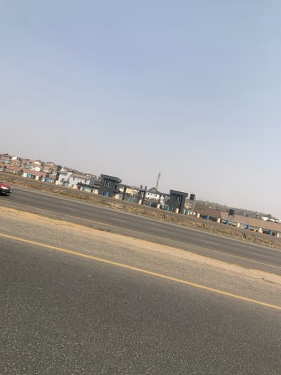 The scene depicts a wide, multi-lane paved road under a clear, hazy sky, likely in the Aso Rock Estate area of Aso, Nigeria. In the immediate foreground, a dark asphalt road surface with a solid yellow line on its right edge and a dashed white line separating lanes is visible. A dirt median strip runs parallel to this road, separating it from another multi-lane paved road in the midground. A single, blurred red vehicle is partially visible moving rightward on the far-left lane of the foreground road.

Beyond the second road, several modern, dark-colored architectural structures, resembling gatehouses or entrance facilities, are present. A prominent white sign above the central structure clearly reads "ASO ROCK ESTATE". To the right and behind these structures, a tall, grey metallic cellular tower or mast stands. The background is dominated by numerous residential buildings with light-colored roofs, scattered across an elevated, urbanized landscape. No individuals or specific interactions between people are discernible. The environment is bright and appears to be daytime.