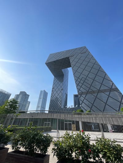 The image shows a daytime view of the China Central Television (CCTV) Headquarters in the Chaoyang District of Beijing, China. The unique loop-like structure of the building is the primary subject. Other skyscrapers are visible in the background. A metal fence and greenery are present in the foreground. The sky is clear and blue.
