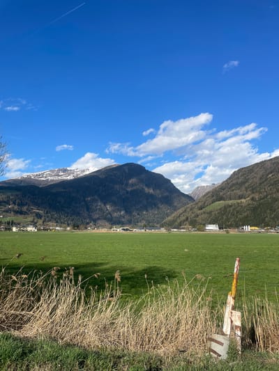 The image depicts a landscape in Sterzing-Vipiteno, Italy, featuring a clear blue sky with scattered cumulus clouds. A mountain range, partially covered in snow, dominates the left side of the frame, while a grassy field occupies the right. Along the edge of the field, there's a patch of dry, brown grass and some green vegetation. In the foreground, near the grass, there's a weathered, yellow and white metal structure. A contrail from a plane can be seen in the upper left quadrant of the image. The environment suggests a rural or suburban area.