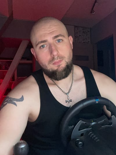 This is a close-up, medium shot portrait of a bald, white male with a goatee and blue eyes, wearing a black tank top and a silver chain with a cross pendant. He has a tattoo on his left arm and is holding a gaming steering wheel with paddle shifters. The background is dimly lit with red lighting, and shelves are visible to the left. The setting is indoors, possibly a home entertainment area. There is no discernible information in the image that would indicate the location as Gliwice, Poland.