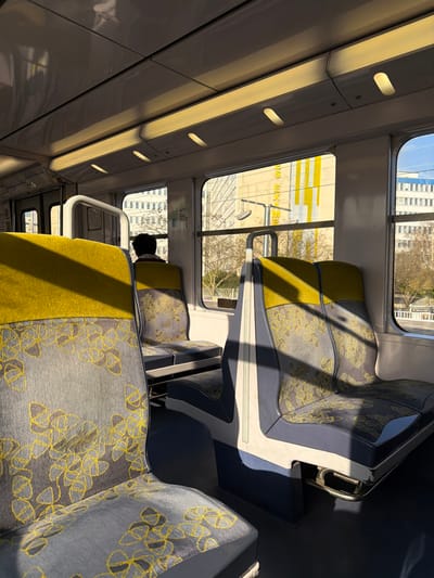 The image displays the interior of a public transport vehicle, likely a train or tram, with an exterior urban view indicating the vicinity of Arcueil, France. The vehicle's interior features rows of empty seats upholstered with a two-tone design: the upper backrests are solid chartreuse yellow, while the lower backrests and seat cushions are grey with a repeating yellow floral or geometric pattern. The seat frames are light grey and dark blue. The floor is dark blue, and the white ceiling contains recessed oval-shaped lighting fixtures. Strong natural sunlight enters from the right, casting defined shadows across the seats and floor.
One individual is present in the middle-ground, seated with their back to the viewer, facing the windows. The back of their head, featuring dark hair, is visible, along with the shoulders of a dark garment that includes a reddish collar or trim.
Through the windows, a prominent, light-colored modern building with distinctive vertical yellow panels is visible. This structure is identified as the ENS Paris-Saclay building, with partial text reading "ES EX..." discernible on its facade. Additional multi-story buildings and bare deciduous trees are visible in the background under a clear blue sky. A utility pole is also present in the exterior scene. The environment is illuminated by bright natural daylight.