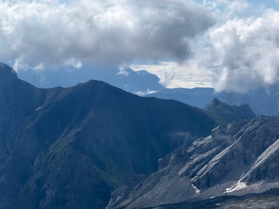 This image captures a view of the Alps, likely taken from a high vantage point. The foreground features rugged, rocky mountain slopes with patches of snow. Further in the distance, several layers of mountain ranges recede into a hazy blue, with lush green vegetation visible on some of the lower slopes. The sky is partly cloudy, with large, dramatic clouds casting shadows over the mountains. The lighting suggests it is daytime, possibly late afternoon given the shadows. There are no people or man-made structures visible in the image. The location is identified as Wildermieming, Austria, known for its alpine landscape.