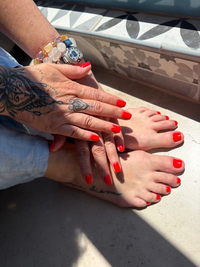 The image shows a close-up of a person's hands and feet, both adorned with bright red nail polish. The person is wearing a light blue garment at the bottom of the frame. On their left wrist, there is a silver watch and a bracelet made of colorful beads, including clear, yellow, and purple stones. The person also has intricate black tattoos on their hands and forearm, one resembling a mandala pattern and another with script.
The subjects are positioned on a textured surface that appears to be a floor or patio, with a shadow cast across it, suggesting it is daytime. The background includes a tiled wall with a decorative pattern. The lighting indicates it is a sunny day, possibly late afternoon due to the angle of the shadows. The overall mood is relaxed and personal, focusing on self-care and adornment. No discernible text other than script tattoos is visible. The location context is Ashkelon, Israel.