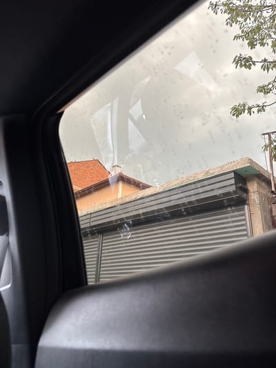 The image is a close-up, slightly angled shot looking upwards through a sunroof. The sunroof glass has water droplets on it, creating a somewhat distorted view of the surroundings. In the upper left quadrant, branches with green leaves hang into the frame, suggesting an outdoor setting with foliage.
The main visible scene through the sunroof depicts the exterior of buildings. To the right, a section of a building with a gray, ribbed metal door or shutter is prominent. Above this shutter, a concrete structure with some exposed brick is visible. To the left of the metal shutter, a building with a terracotta tile roof is partially visible, appearing to be older construction. The sky is overcast and appears bright, suggesting daytime, possibly a cloudy or rainy day given the water on the sunroof.
The scene is framed by the dark interior of a vehicle, likely a car, with the edges of the sunroof opening and parts of the car's interior visible in the foreground and along the right side of the image. There is no visible text in the image. The overall impression is a view from inside a vehicle on a day with light rain, looking out at urban architecture. The location is Gotse Delchev, Bulgaria.