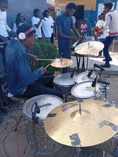 This image captures an outdoor musical performance in Jebbu Miango, Nigeria. In the immediate foreground, a male drummer, wearing a denim jacket, dark trousers, and maroon-banded headphones, is seated on a brown stool, actively playing a five-piece drum kit. He holds wooden drumsticks, poised over a gold Sabian crash/ride cymbal and a tom drum. The drum kit features a snare, two rack toms, a floor tom, and a bass drum, some with a distinctive light blue patterned finish, and three Sabian-branded cymbals, two of which show dark tape patches. Two microphones are clipped to the drums.
To the drummer's left, a male in a white long-sleeved shirt plays a keyboard. To the drummer's right, a second male keyboardist, dressed in a dark blue traditional tunic, operates another keyboard on a stand. Further to the right, a male vocalist, wearing a white vest with red horizontal stripes and dark trousers, sings into a handheld microphone while simultaneously playing a brown four-string bass guitar. Behind these individuals, several other vocalists, including two women and a man in white t-shirts, are also singing into handheld microphones.
The background consists of a light-colored building, transitioning to a section painted blue on the right, where white signs display partial text, including "OPEN," "RUN," and "WITH." A dense green bush is centrally located behind the drum kit. The ground is unpaved earth, with multiple audio cables visible. A child in a pink shirt is discernible in the distant background near the blue building. The scene is illuminated by natural daylight.