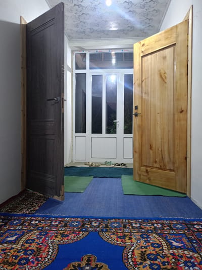 The image depicts an interior hallway with two open doors. The door on the left is dark wood with horizontal decorative grooves and a silver handle. The door on the right is light-colored wood, also with horizontal decorative elements and a black handle. Both doors are ajar, revealing an exit area with a white framed window and door combination.
On the floor, there are several rugs. A large, ornate rug with intricate patterns in red, blue, yellow, and black is in the foreground. Behind it, a solid blue rug is visible, followed by two smaller rugs, one green and one dark green. A pair of light-colored sandals and a pair of dark sandals are placed on the dark green rug near the exit.
The walls are painted white, and the ceiling has a decorative pattern, possibly wallpaper or textured tiles, in white and gray. The lighting suggests it is either early morning or late evening, with some artificial light sources illuminating the space. The overall impression is of a domestic entryway in a building located in Panjakent, Tajikistan. No people are present in the scene.