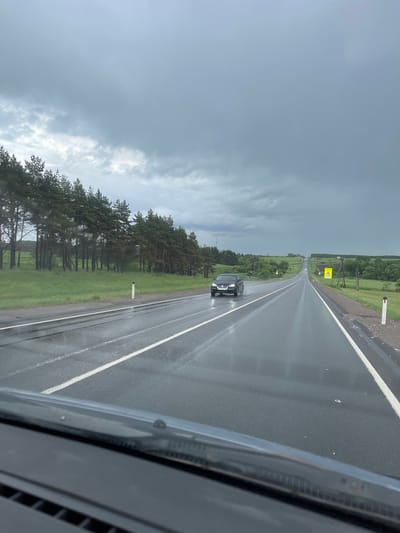 The image depicts a car traveling on a wet, asphalt road in the vicinity of Kokino, Russia. The road is marked with a white lane divider. A black car is visible ahead on the same lane, moving in the same direction. Trees line the left side of the road, bordering a green grassy area. A road sign is visible further down the road. The sky is overcast with dark, grey clouds, suggesting recent or ongoing rain. The image is taken from inside a vehicle, with parts of the car's interior visible in the foreground.