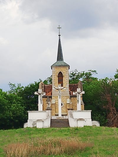 This is a medium shot, eye-level, front-facing photograph of a small, old, brick church with a pointed steeple topped with a cross. The church is built with light-colored bricks and has a tall, arched window. In front of the church are three large crucifixes, each with a statue of Jesus on it. There are also two statues of men on pedestals. A set of stairs leads up to the church entrance. The church and its statues are surrounded by lush green trees, and the foreground is a grassy hill with dry grass and some wildflowers. The sky is overcast with gray clouds. The scene appears to be tranquil and somewhat somber. The overall impression is of a historic religious site set in a natural landscape. There is no visible text in the image.