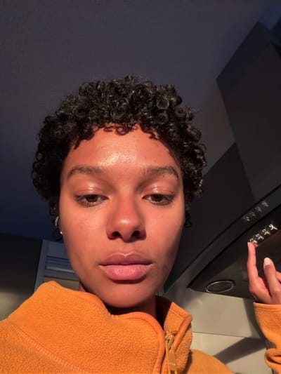 A close-up, angled shot shows a person with brown skin and curly black hair. They are wearing an orange fleece hoodie. Their right index finger is pointing towards a series of metal switches, possibly on a kitchen appliance. The background is a mix of dark gray and off-white surfaces. The location in Champs-sur-Marne, France is not evident from the image.
