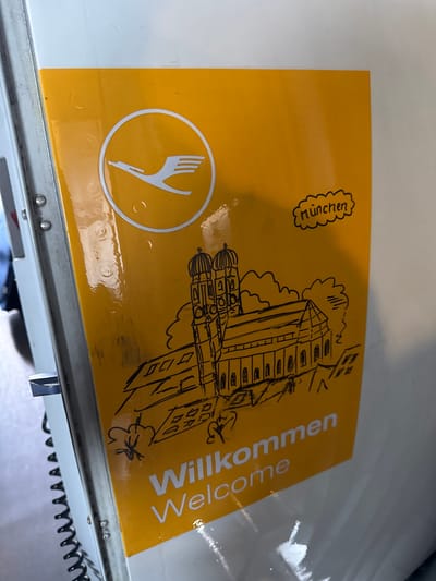 The image shows a yellow welcome card from Lufthansa airlines on a white surface, likely the interior of an aircraft. The card features the Lufthansa logo, a drawing of a building identified as being in Munich (written as "München" in German), and the words "Willkommen Welcome" in white. The image is taken in a slightly angled, close-up perspective, revealing some minor water droplets on the card's surface. The bottom edge of the image includes part of an electrical cord. The presence of the "München" logo suggests the location is likely the airport in Oberding, Germany.