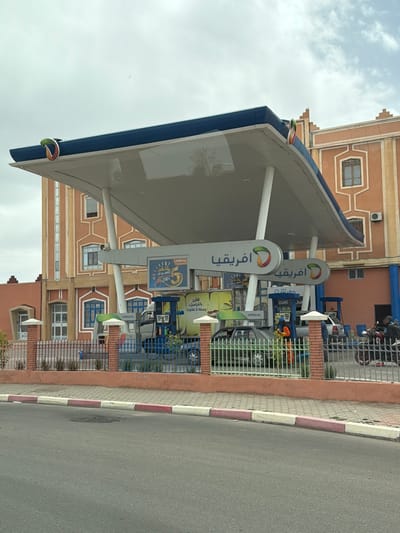 This is an exterior shot of an Afriquia gas station located in Ouarzazate, Morocco. The gas station has a large white canopy with a blue trim, supported by white pillars. Signage with the Afriquia logo and Arabic script is prominently displayed. The gas pumps are visible behind the pillars, along with advertisements and branding. A motorcycle is parked behind a protective fence, while vehicles can be seen lined up at the pumps. The building behind the gas station is light brown, with distinct architectural detailing. The image also features a segment of a road with a curb painted in alternating white and red sections. The sky is overcast.