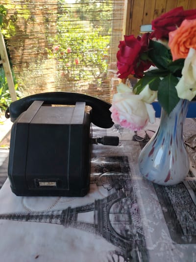 The image shows a close-up view of a vintage black telephone on a table covered with a patterned tablecloth. The tablecloth features a depiction of the Eiffel Tower. Beside the telephone, a vase filled with roses of various colors including red, orange, and white is placed. In the background, a woven bamboo blind is visible, with greenery and hints of red flowers seen through it, suggesting an outdoor or semi-outdoor setting. The scene is illuminated by natural light, casting shadows on the objects. The overall impression is one of a still life arrangement. The location is identified as Kozare, Bulgaria.