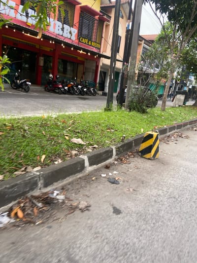 The image shows a street in Margaasih, Indonesia, located at coordinates -6.968 latitude and 107.553 longitude. The street is paved and lined with a curb and a grassy area. Several motorcycles are parked along the curb, in front of a commercial building with a sign that reads "CAFE & RESTO." The building's facade is painted in shades of yellow and red, and there is a second sign that reads "LAMIRRY" in large, stylized letters. There is a yellow and black traffic cone on the street near the curb. The ground is littered with debris. Several trees are visible in the background.