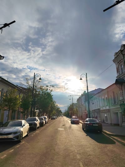 The image depicts a street scene in Rybinsk, Russia. The sun is low in the sky, casting long shadows and illuminating the road with a warm, golden light. The sky is partially overcast with dramatic clouds, and the sun is breaking through, creating a lens flare effect. The street is lined with buildings on both sides, some appearing to be old, with ornate architecture. Trees are planted along the sidewalks, and several streetlights are visible. Multiple cars are parked along the right side of the road and are also driving on the street. One car in the foreground on the left is a silver Kia with a license plate visible. On the right side of the street, a dark sedan with a license plate is also visible. A pedestrian can be seen in the distance walking on the sidewalk. Overhead, there are wires and decorative banners stretched across the street. The overall atmosphere suggests a late afternoon or early evening in a city with a mix of historic and modern elements.
