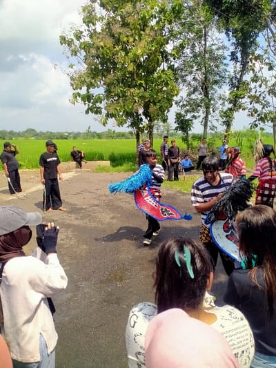 A group of people are gathered in an open field in Kecamatan Kedawung, Indonesia (latitude: -7.52, longitude: 111.027).  The individuals are positioned on a paved pathway adjacent to a rice paddy field.  One man stands to the left with a long stick in his hand, another is seated on a bench nearby.  The people are mostly wearing casual clothing. There are two men performing a traditional dance with blue and red fabric. One person is seated on the pathway to the left, watching the performance.  To the right of the dancers are several people watching the performance, including one woman who is filming the scene.  The background of the scene features a field of green rice paddy with large trees lining the edge of the field. The sky is cloudy.