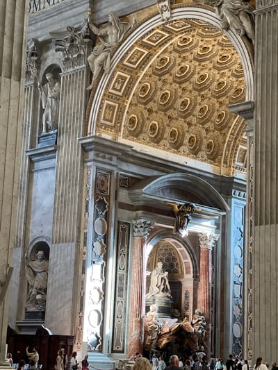 The image depicts a detailed interior view of St. Peter's Basilica, located in Città del Vaticano, Vatican City. The perspective is directed towards the upper left and center-right, showcasing a section of the opulent architecture and statuary.
In the upper central portion, a large arched vault is visible, extensively decorated with an array of octagonal coffers, each containing an ornate golden rosette or floral motif. Surrounding these coffers are smaller rectangular and square decorative panels. An inscription in large capital letters, partially visible at the top left, reads "OMNIVM".
On the far left, a tall, light-colored marble pilaster with vertical fluting runs from top to bottom. Within a niche embedded in this wall stands a full-length marble statue of a bearded male figure, robed and holding a staff, with a smaller animal (possibly a lamb) at his feet. Below this statue, a dark plaque bears the partially legible inscription "S. GYLVLIUS ABBAS". Further down the wall, another niche contains a robed figure holding a torch aloft. Wooden architectural elements, possibly parts of confessionals or benches, are visible at the base of the left wall.
On the right side of the image, a massive, fluted, round marble column rises vertically. Adjacent to it, a wall section is intricately decorated with large, rectangular panels of multi-colored marble intarsia, featuring geometric and curvilinear patterns in shades of blue, brown, black, and white.
Towards the lower central area, a significant sculptural monument, identifiable as a Papal tomb, is prominent. It features a central seated figure (likely a Pope) atop a pedestal, surrounded by dynamic, draped allegorical figures and flowing marble elements in reddish-brown and white hues. A plaque below the central figure is partially visible with the name "ALEXANDER". Above this monument, an ornate, gilded eagle or bird-like sculpture is mounted on an arch, reflecting a strong highlight.
Numerous individuals, appearing to be tourists or visitors, are present in the lower foreground and mid-ground. They are casually dressed, standing, walking, and observing the surroundings. Some individuals are holding smartphones, suggesting they are taking photographs or videos. Strong natural light enters from the right side, creating pronounced highlights and shadows that emphasize the textures and depths of the marble and gilded decorations.