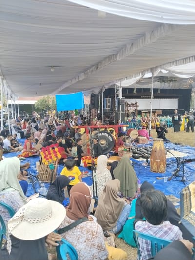 A wide-angle, eye-level shot captures an outdoor performance or gathering under a large white tent. The tent's draped fabric creates a sense of enclosure, with some lights visible. The ground is covered in a bright blue tarp, on which a group of people are seated, some on chairs and others directly on the tarp.

In the foreground, several individuals are seen from behind, facing a performance area. One person wears a wide-brimmed straw hat decorated with a polka-dot scarf. Another wears a patterned headscarf. A young child in a yellow t-shirt sits on the tarp.

The central focus of the scene is a musical ensemble. Several people are seated on the blue tarp around various musical instruments, including what appear to be gamelan instruments such as gongs and metallophones. A large drum is prominently displayed, as is a large gong in the middle ground. The musicians are dressed in traditional attire, with some wearing red and yellow sashes.

In the background, a stage is set up, indicated by a white screen and a dark backdrop. Several performers are seated on chairs on the stage, dressed in elaborate costumes. A few individuals are standing near the stage, including someone holding a camera, suggesting a recording of the event. The overall atmosphere appears to be a cultural performance or festival. The location is identified as Plosorejo, Indonesia.