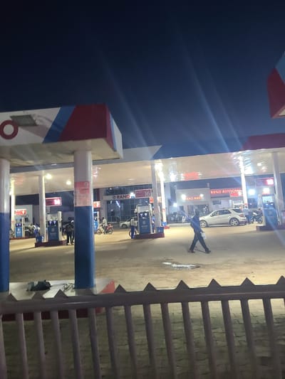 This image depicts a night scene at a RANO Autocare and RANO Mart gas station in Nyanya, Nigeria. Several people are visible, including a person standing in the middle of the forecourt, seemingly posing with arms and legs spread wide. Other individuals are seen near gas pumps and in the background, suggesting a busy evening at the station.
Objects present include cars, motorcycles, and multiple gas pumps, illuminated by the station's lights. The RANO Autocare and RANO Mart signs are clearly visible on the building, indicating the services offered. The ground appears wet in places, possibly from recent rain or cleaning, and the overall lighting suggests it is nighttime. The presence of a white car and motorcycles indicates vehicles are either refueling or parked. A metal fence with pointed posts is prominent on the right side of the image, bordering the gas station's area. The sky is dark, consistent with a night setting. The scene captures the everyday operations of a gas station in Nigeria during the evening.