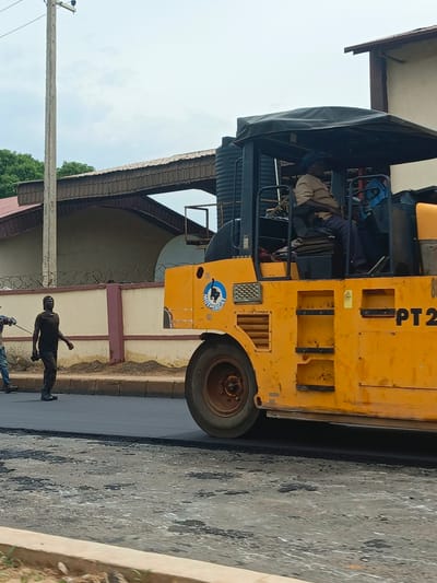 In Tudun Nupawa, Nigeria, a bright yellow road roller is compacting fresh asphalt on a street. A man wearing a tan shirt and dark pants operates the roller from its cabin. In the foreground, another man in a dark shirt and pants walks away from the roller, holding what appears to be a tool in his right hand. The street is lined with a low wall topped with barbed wire, behind which are buildings with reddish-brown tiled roofs and a beige facade. A utility pole stands to the left of the frame, supporting power lines. The sky is overcast.
