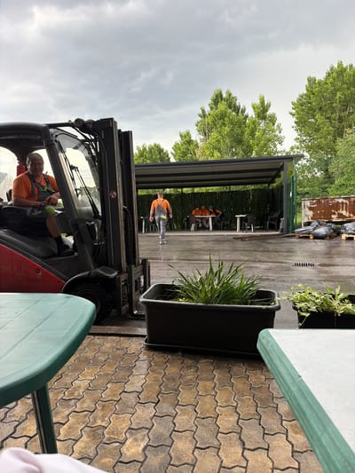 The image depicts an outdoor scene, possibly a recycling or waste management facility, under a cloudy sky. In the foreground, a man wearing an orange shirt and grey overalls is seated in the driver's seat of a red and grey forklift. He is looking towards the camera. To the right of the forklift, a large black planter box contains green, grass-like plants. In the background, under a covered shelter, several people in orange shirts are seated around a table. Further to the right, there are stacks of black garbage bags on wooden pallets and a rusty corrugated metal structure. The ground is paved with interlocking bricks and appears wet. Two green tables are partially visible in the foreground, one on the left and one on the right. The location is identified as Lozen, Bulgaria.