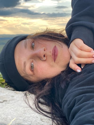 This is a selfie taken by a person with fair skin, green eyes, and dark brown hair. They are wearing a black hoodie and have a silver ring on their nose. The person is lying down, with their head tilted and their face close to the camera. Their hand is resting on their forehead, and their fingernails are painted white. The background shows a cloudy sky with golden light, suggesting it is either sunrise or sunset. The setting appears to be outdoors, possibly on a rocky surface. The overall mood seems relaxed and contemplative.